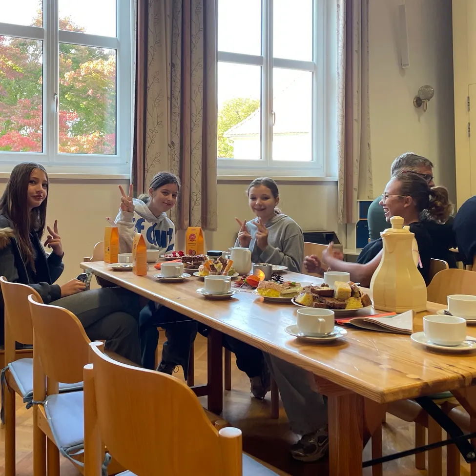 Eine Gruppe von Menschen sitzt um einen Tisch mit Tassen, Untertassen und verschiedenen Speisen. Der Tisch ist mit Tellern, Tassen und Untertassen besetzt. Der Raum hat große Fenster mit Vorhängen.