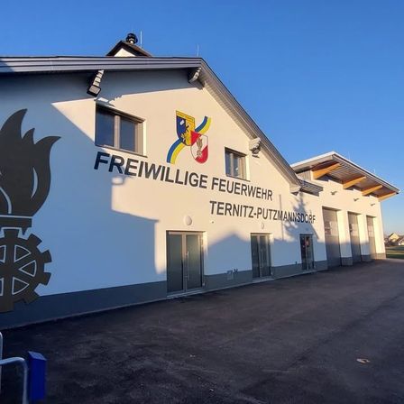 Ein weißes Gebäude zeigt den Text 'Freiwillige Feuerwehr Ternitz-Putzmannsdorf'. Es hat zwei Haupttüren, zwei Fenster und ein Logo. Der Himmel ist klar.
