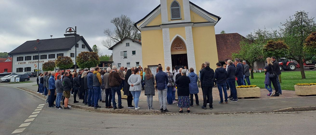 Eine Menschenmenge steht vor einer Kirche mit einer Uhr auf dem Turm. Die Kirche hat ein weiß-gelbes Farbschema.