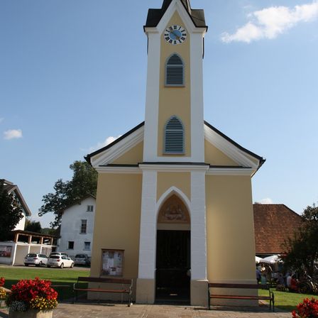 Eine kleine Kirche mit einem spitzen Turm, einer Uhr und Bogenfenstern, steht gegen einen blauen Himmel mit weißen Wolken.