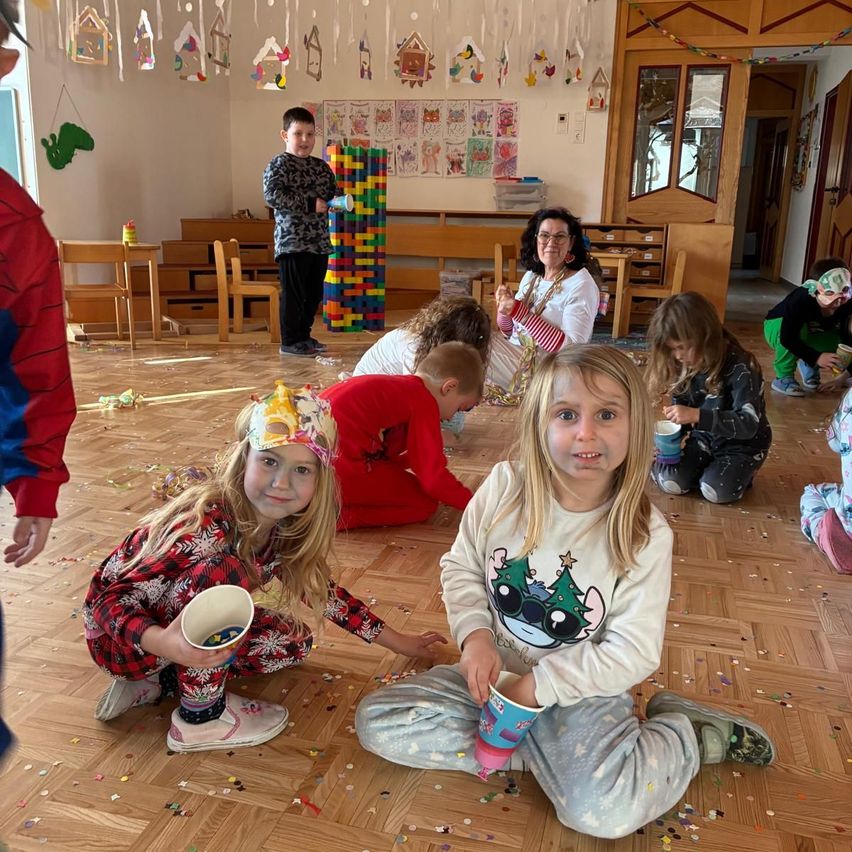 Kinder spielen auf dem Boden in einem Klassenzimmer. Einige sitzen, andere stehen. Ein Kind hält einen Becher mit Konfetti. Eine Frau steht in der Nähe eines Stapels bunter Bausteine.