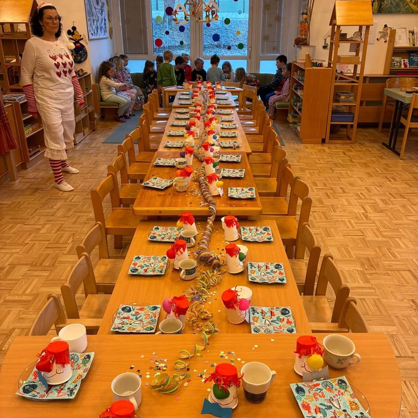 Ein langer Esstisch für eine festliche Veranstaltung, geschmückt mit bunten Dekorationen, Tellern und Tassen. Eine Frau steht an einem Ende, und Kinder sitzen am anderen, in der Nähe eines Fensters mit Luftballons und einem Bücherregal.