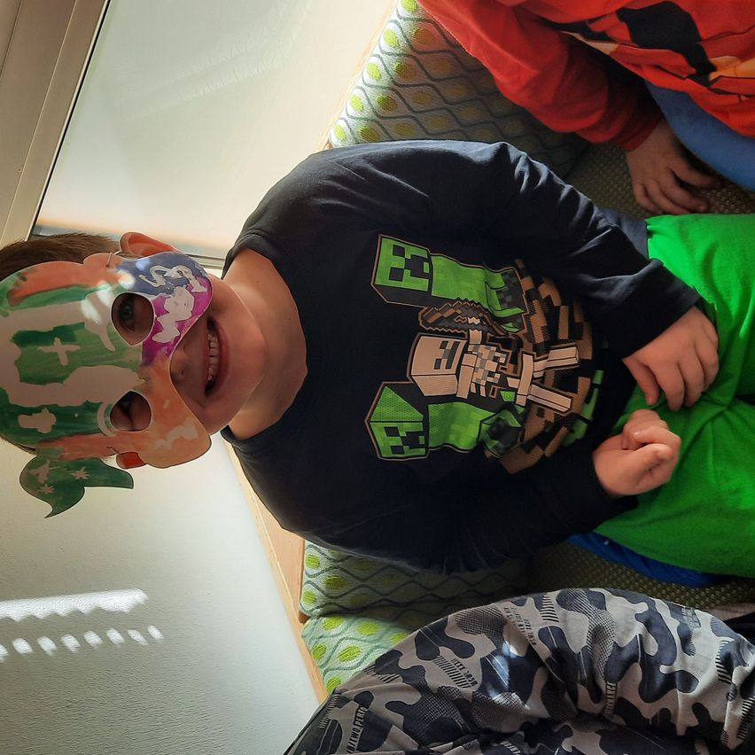 Ein Kind mit einer farbenfrohen Maske und einem lächelnden Gesicht sitzt auf einer Couch und schaut nach vorne.