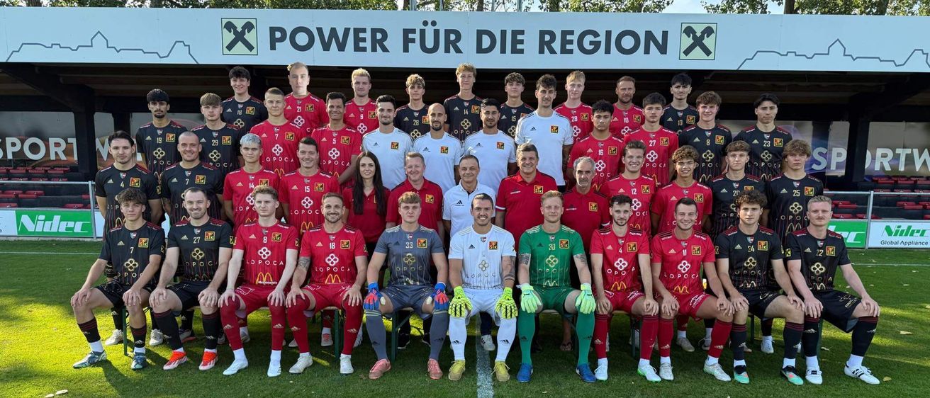 Eine Fußballmannschaft steht auf einem Feld mit einem Banner, das 'Power für die Region' besagt. Die Mannschaft trägt rote und weiße Uniformen, und es gibt zwei Frauen in der Mitte. Die Mannschaft posiert für ein Foto.