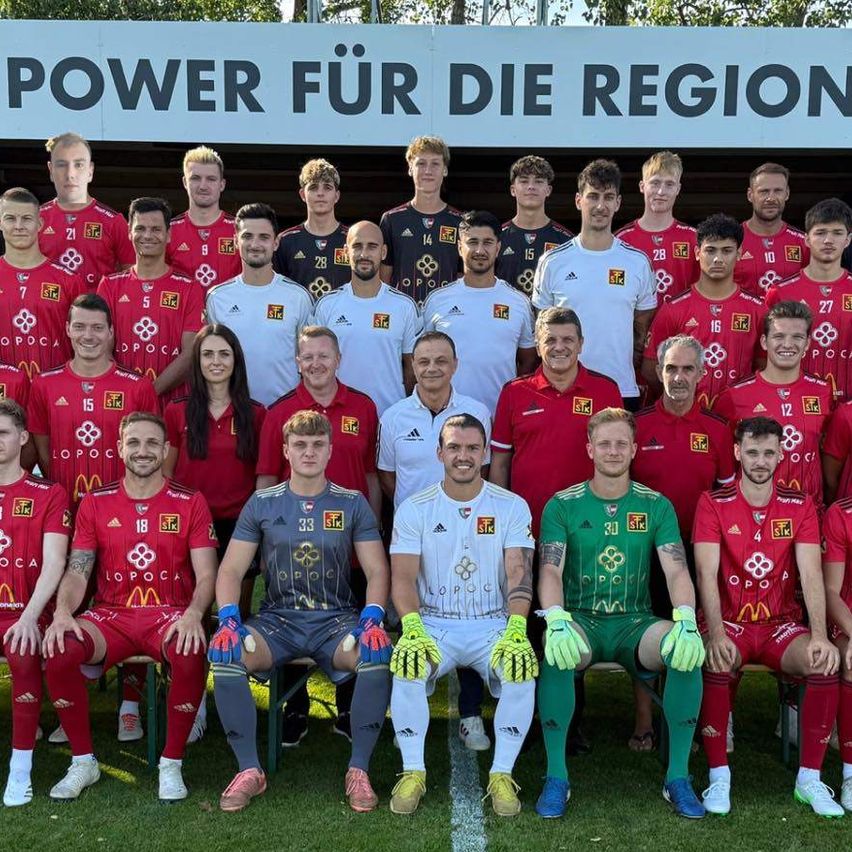 Eine Fußballmannschaft steht auf einem Feld mit einem Banner, das 'Power für die Region' besagt. Die Mannschaft trägt rote und weiße Uniformen, und es gibt zwei Frauen in der Mitte. Die Mannschaft posiert für ein Foto.