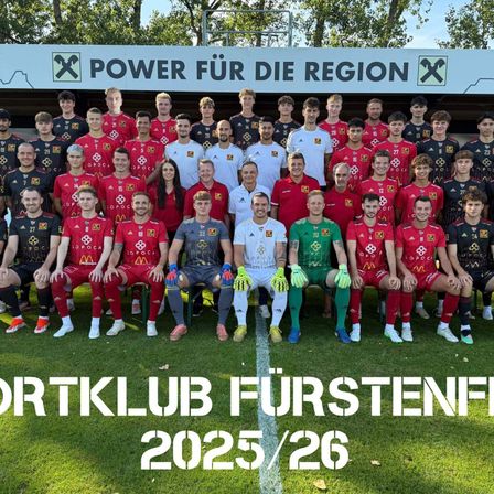 Eine Fußballmannschaft posiert für ein Foto auf einem Feld, in roten und weißen Trikots gekleidet. Ein Banner hinter ihnen trägt den Text 'Power für die Region'. Die Mannschaft ist als 'Sportklub Furstenfeld 2025/26' beschriftet.