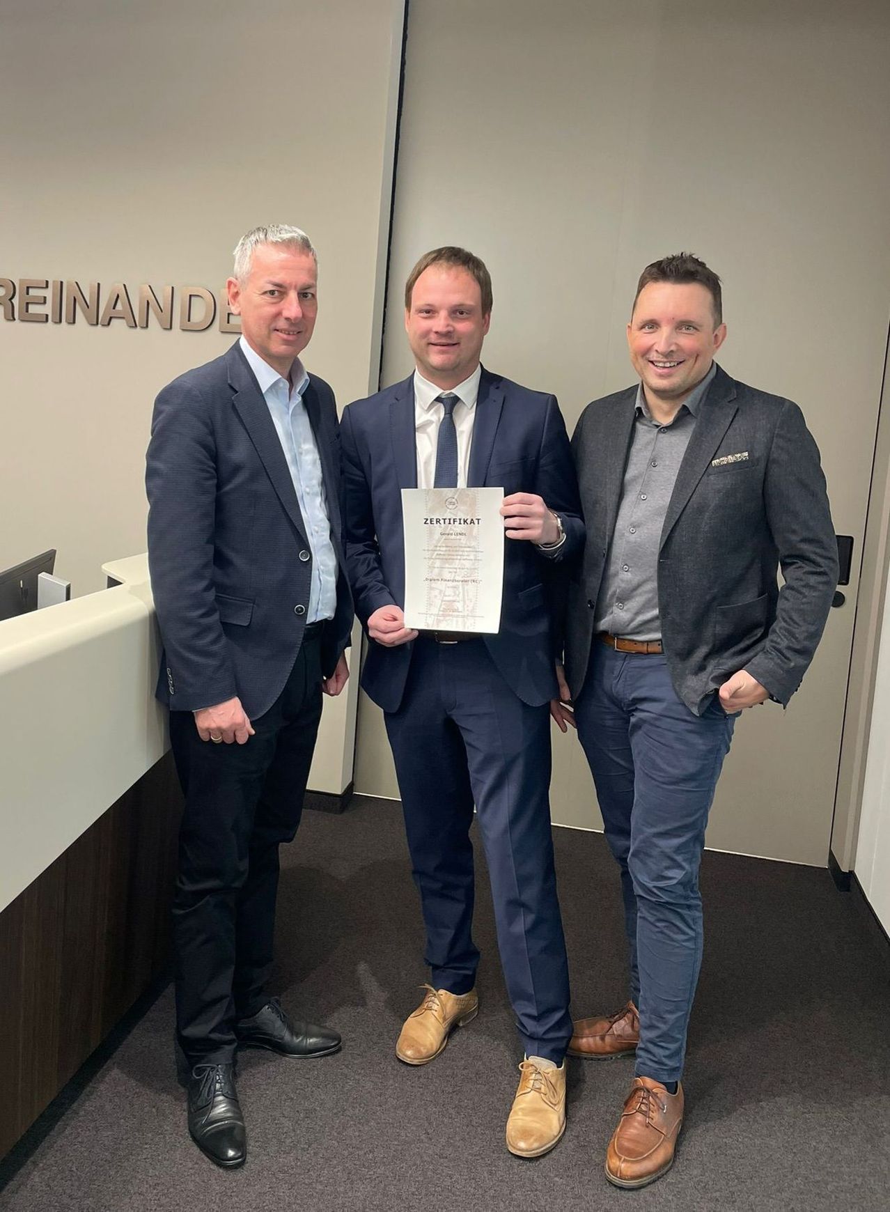 Drei Männer in formeller Kleidung stehen in einem Büro, einer hält ein Zertifikat. Sie posieren lächelnd für ein Foto. Hinter ihnen befindet sich eine Wand mit dem Wort 'Reinand'.