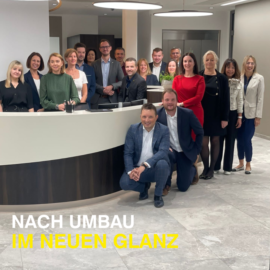 Eine Gruppe von Menschen in formeller Kleidung, die hinter einer Rezeptionstheke mit einer Pflanze stehen und sitzen, lächeln und für ein Foto posieren. Das Foto hat einen Wasserzeichen mit dem Text 'NACH UMBau IM NEUEN GLANZ'.