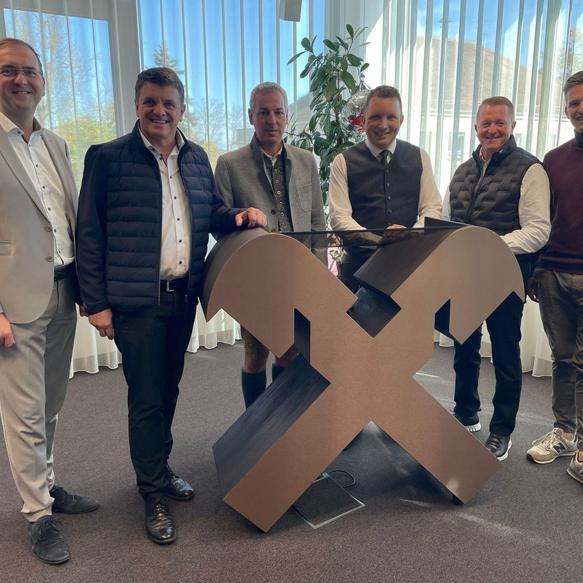 Fünf Männer stehen um ein großes X-förmiges Objekt in einem Büro, lächeln und posieren für ein Foto. Hinter ihnen sind Fenster und eine Pflanze.