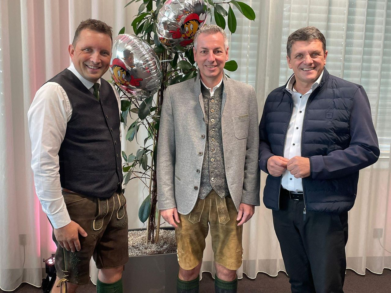 Drei Männer posieren für ein Foto, zwei in traditioneller deutscher Kleidung, einer in einer Jacke, hinter einer großen Pflanze mit Luftballons.