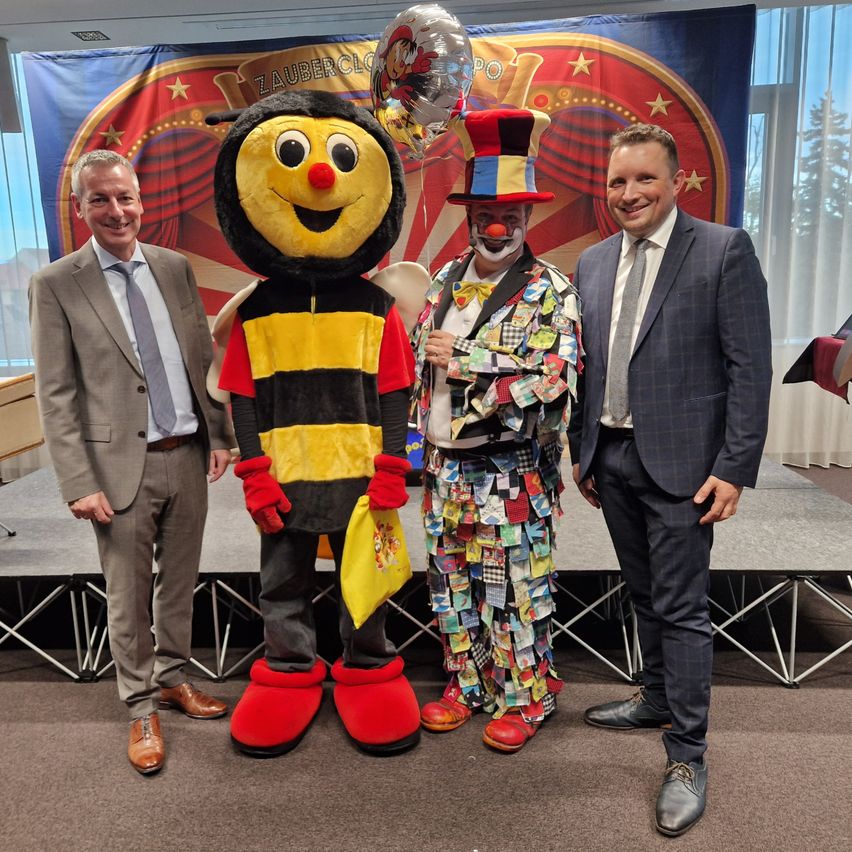 Drei Männer stehen auf einer Bühne. Einer trägt ein Bienen-Mascot-Kostüm, einer ein Clown-Kostüm und der dritte einen Anzug. Ein Banner mit den Worten 'Zaubercircus' ist hinter ihnen.