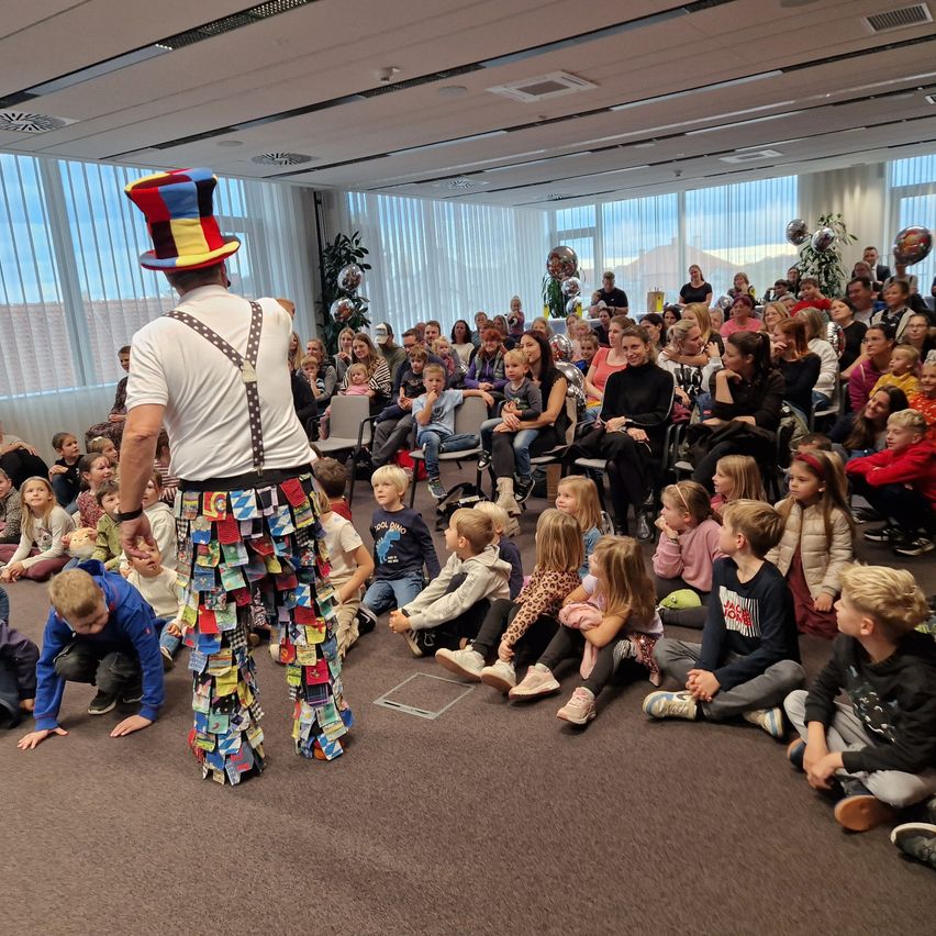 Ein bunt gekleideter Mann unterhält ein großes Publikum aus Kindern und Erwachsenen in einem Konferenzraum.