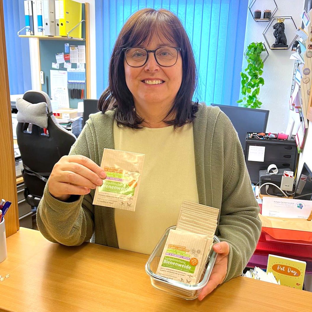 Eine Frau mit Brille und einem grünen Cardigan hält einen Beutel mit Bienenpollen und einen Behälter mit mehr Bienenpollen in einem Büro.