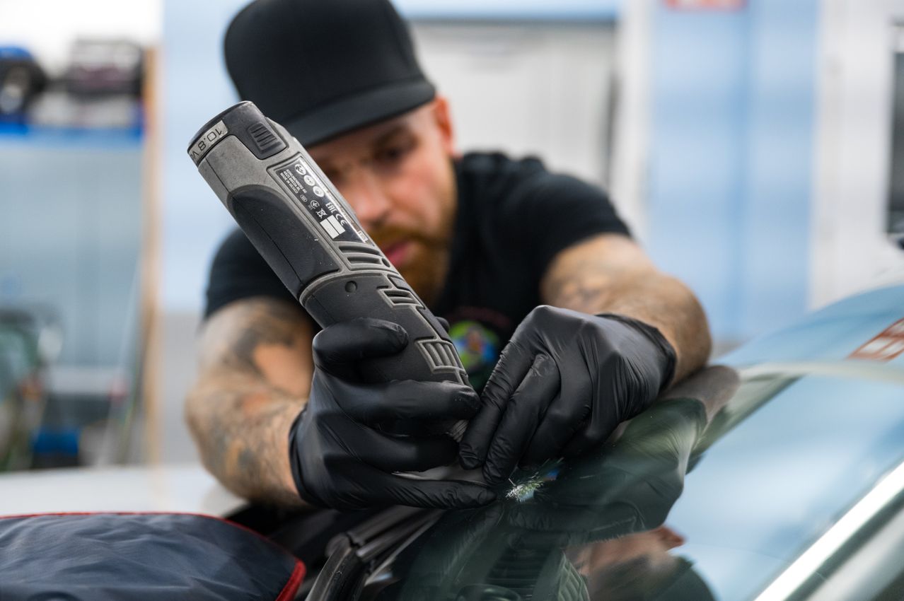 Ein Mann mit schwarzem Hut und schwarzen Handschuhen benutzt ein Werkzeug auf einer Windschutzscheibe. Er scheint an der Windschutzscheibe des Autos zu arbeiten.