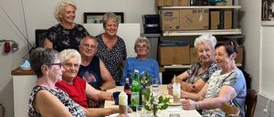 Sieben ältere Erwachsene versammeln sich um einen Tisch, lächeln und posieren für ein Foto. Sie haben Getränke und Essen vor sich. Dahinter sind Regale mit Kisten und Lagerboxen zu sehen.