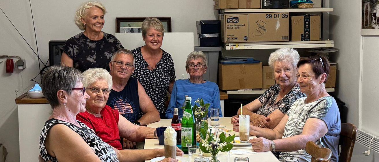 Sieben ältere Erwachsene versammeln sich um einen Tisch, lächeln und posieren für ein Foto. Sie haben Getränke und Essen vor sich. Dahinter sind Regale mit Kisten und Lagerboxen zu sehen.