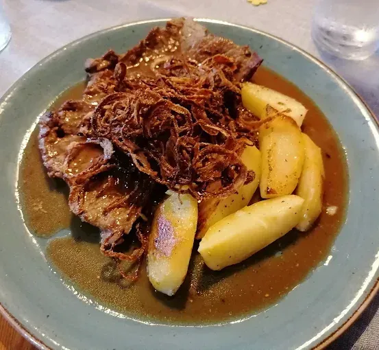 Ein Teller mit einem Fleischgericht, bestehend aus Kartoffelscheiben und einer braunen Soße, auf einem Holztisch.