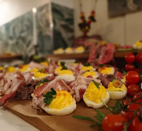 Ein Holzbrett zeigt eine Auswahl an gefüllten Eiern, Fleischscheiben und Kirschtomaten. Die Eier sind mit gelben Eigelben und Kräutern garniert. Das Fleisch ist dünn geschnitten und neben den Eiern angeordnet. Die Kirschtomaten sind frisch und leuchtend.