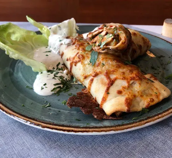 Ein Teller mit zwei aufgerollten Speisen, eine mit geschmolzenem Käse und Kräutern, die andere mit einer cremigen Dip-Sauce, begleitet von Salat und einer Scheibe Brot.