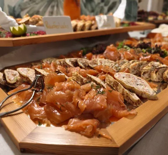 Ein hölzernes Tablett gefüllt mit verschiedenen geräucherten Fisch- und Fleischscheiben, mit Kräutern garniert, begleitet von einem Paar Zangen.