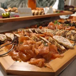 Ein hölzernes Tablett gefüllt mit verschiedenen geräucherten Fisch- und Fleischscheiben, mit Kräutern garniert, begleitet von einem Paar Zangen.