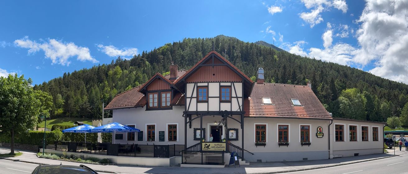 Das Gasthaus zum Osterreich ist ein traditionelles Gebäude mit einem roten Dach, gelegen am Fuße eines grünen Berges. Der Eingang ist mit einem Banner versehen und eine Frau steht neben der Tür.