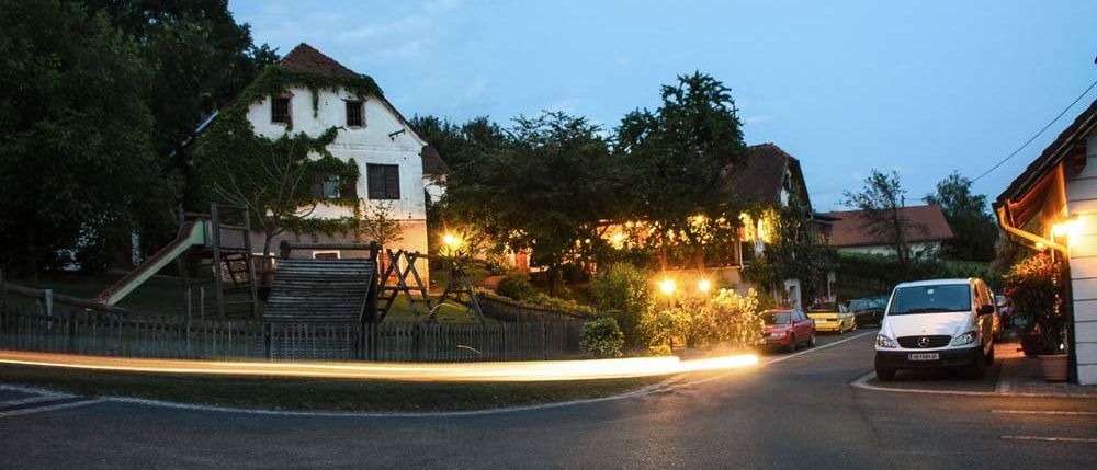 Ein Wohngebiet mit einem Haus, nachts beleuchtet mit einer Schiebetür und einer Rutsche. Bäume und eine Schaukel sind im Garten. Ein weißes Auto ist auf der Straße geparkt.
