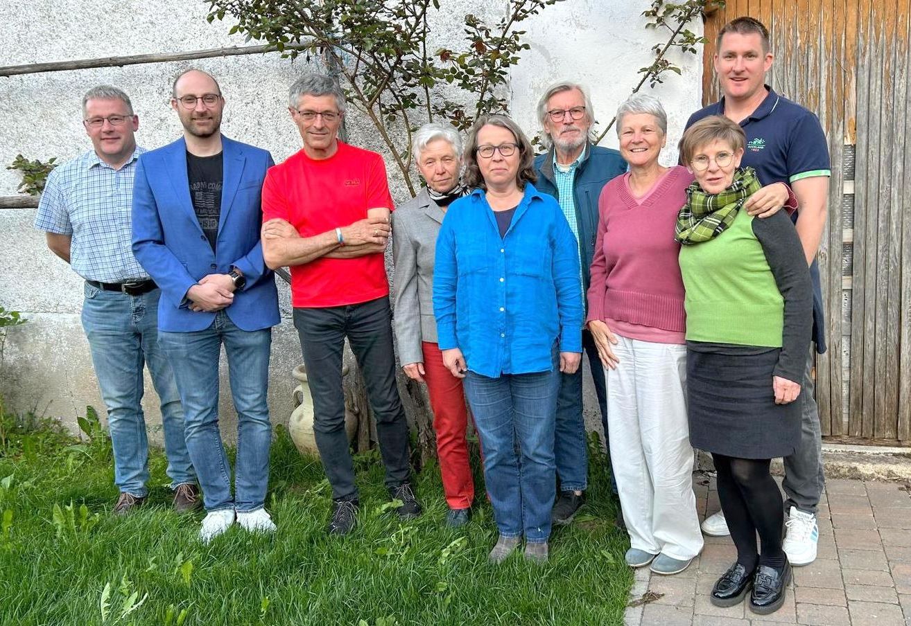Eine Gruppe von Erwachsenen steht draußen in der Nähe einer Wand mit einem Baum. Sie alle lächeln und scheinen für ein Foto zu posieren.