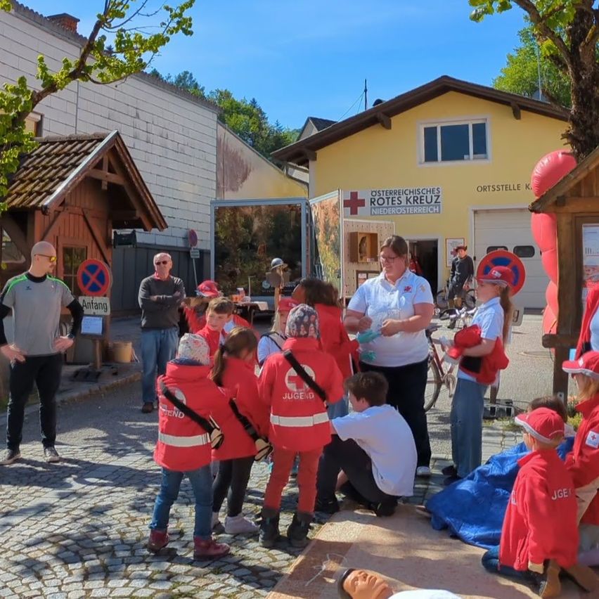Eine Gruppe von Kindern und Erwachsenen in roten Mänteln versammelt sich vor einem Gebäude mit einem roten Kreuzschild. Eine Frau in einem weißen Hemd spricht zu den Kindern.