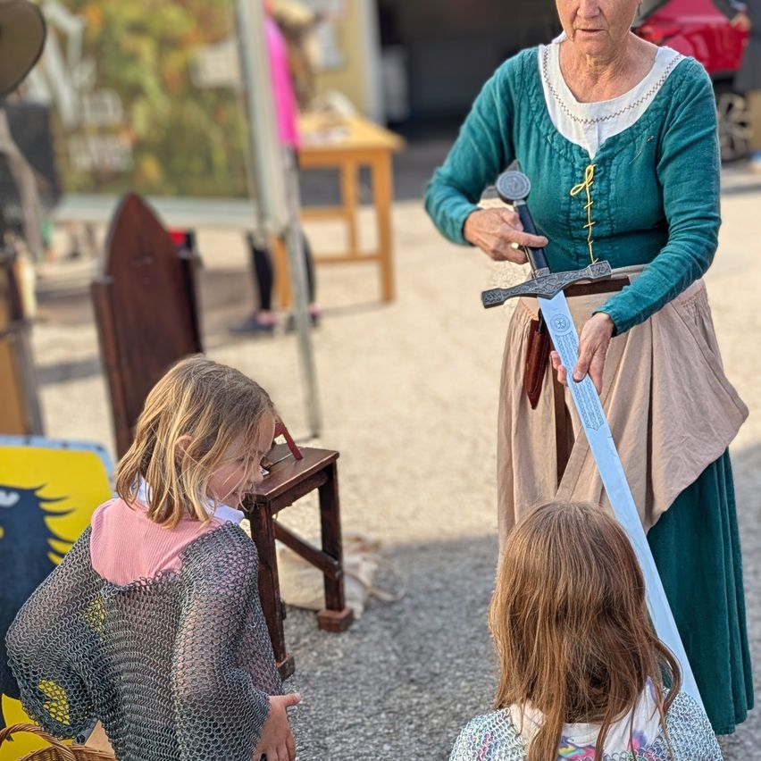 Eine ältere Frau in mittelalterlicher Kleidung hält ein Schwert und demonstriert es zwei jungen Mädchen bei einer historischen Nachstellung.