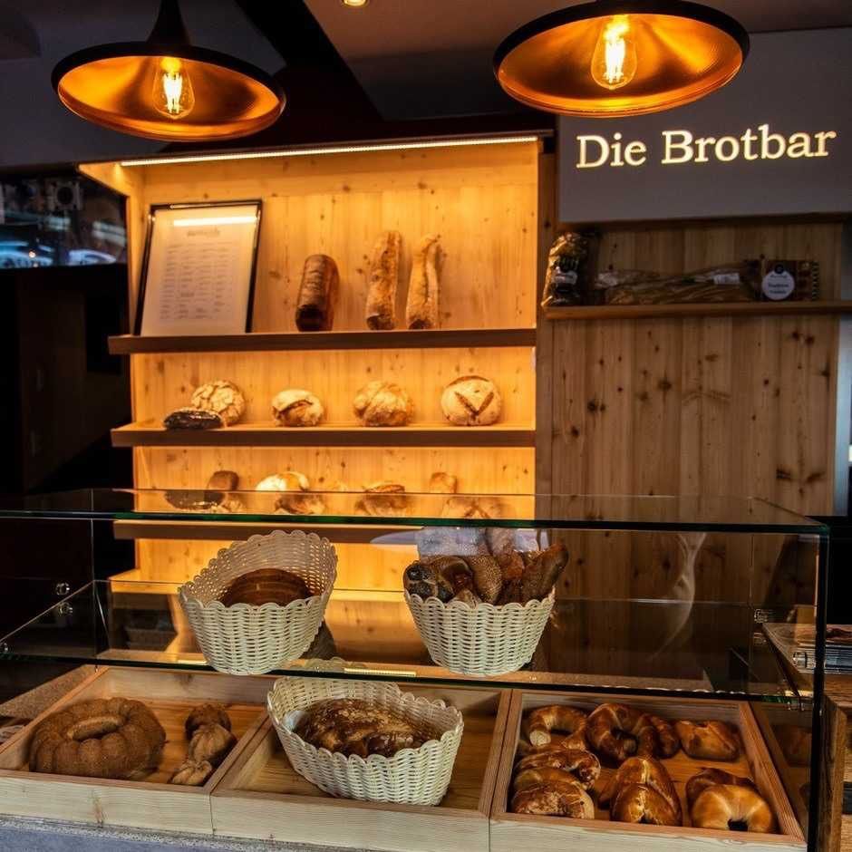 Ein Bäckerei-Display mit einem Schild, das 'Die Brotbar' liest, zeigt verschiedene Brote in Körben und Tabletts, unter warmem Licht von Lampen.