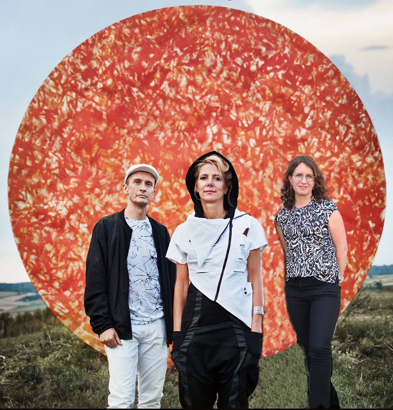 Drei Personen stehen vor einem großen kreisförmigen Gemälde. Der Mann trägt einen Hut und eine schwarze Jacke, die Frau in der Mitte ein weißes Oberteil und die Frau auf der rechten Seite eine Brille und ein gemustertes Oberteil.