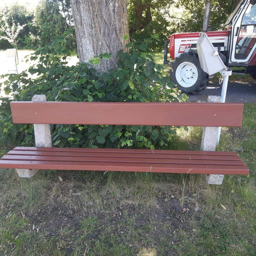 Eine Holzbank befindet sich in einem Grasbereich in der Nähe eines Baumes und eines roten Traktors. Die Bank ist braun mit weißen Akzenten. Hinter der Bank steht ein großer Baum mit grünen Blättern.