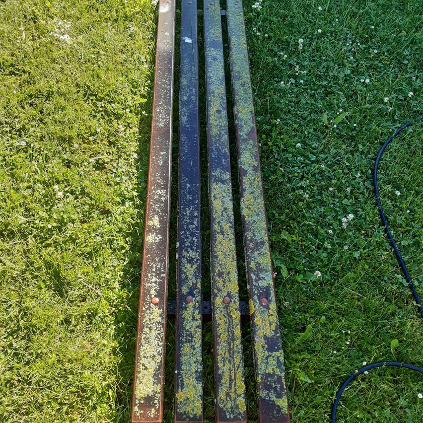 Drei verrostete Metallstangen mit Moos liegen auf einem grünen Rasen mit kleinen weißen Blüten und einem schwarzen Schlauch in der Nähe.