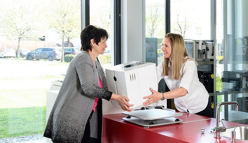 Zwei Frauen in einem Küchenbereich, eine hält ein Gerät, während die andere lächelt und sie ansieht. Hinter ihnen gibt es Glasfenster mit Blick auf geparkte Autos und Bäume.