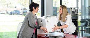 Zwei Frauen in einem Küchenbereich, eine hält ein Gerät, während die andere lächelt und sie ansieht. Hinter ihnen gibt es Glasfenster mit Blick auf geparkte Autos und Bäume.