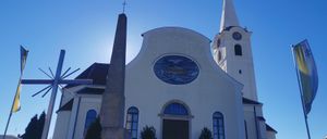 Eine weiße Kirche mit einem hohen Turm und einem blauen runden Fenster. Ein hoher Kreuz und ein Uhrturm sind sichtbar.