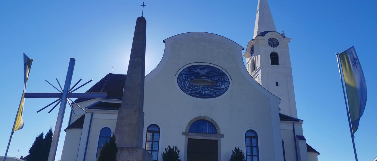 Eine weiße Kirche mit einem hohen Turm und einem blauen runden Fenster. Ein hoher Kreuz und ein Uhrturm sind sichtbar.
