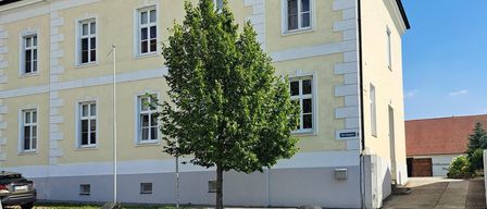 Ein beigefarbenes Gebäude mit mehreren Fenstern steht neben einem Baum, mit einem Auto auf der linken Seite. Das Gebäude hat ein braunes Dach. Ein Bürgersteig befindet sich vor dem Gebäude.