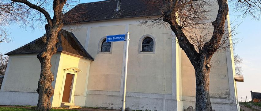 A white church with a brown roof and tower, surrounded by trees, has a blue street sign reading 'Prátal Zistler Platz'. The building has a door and a window.