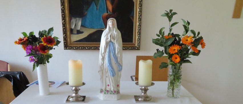 Ein hölzerner Altar, mit weißem Tuch bedeckt, beherbergt eine Statue, zwei Kerzen und zwei Blumenvasen. Ein Gemälde hängt an der Wand dahinter.