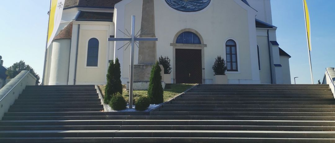 Eine weiße Kirche mit einem spitzen Turm und einem Kreuz darauf, unter einem klaren blauen Himmel. Ein Kreuzmonument steht vor dem Eingang.