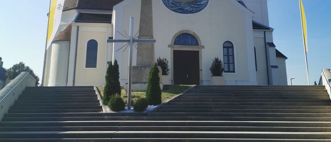Eine weisse Kirche mit einem Kirchturm, einem Kreuz und einer Uhr. Sie hat ein rundes Fenster mit einer Tauben-Design und ein Kreuz-Monument davor.