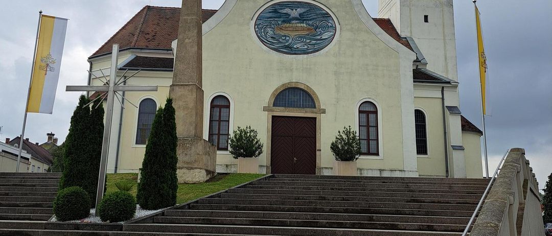 Eine Kirche mit Turm, Uhr und Kreuz oben. Es gibt ein kreisförmiges Buntglasfenster an der Vorderwand. Ein Kreuz und Fahnen befinden sich an den Seiten.