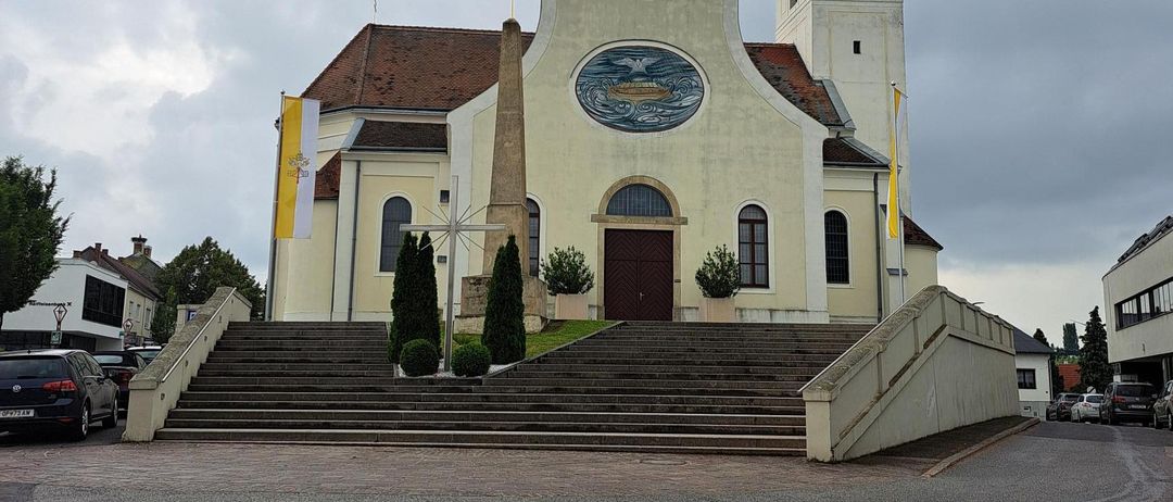 Eine Kirche mit einem Kirchturm und einer Uhr, mit einem kreisförmigen Emblem über dem Eingang. Sie hat Stufen, die zum Eingang führen, mit Fahnen auf beiden Seiten.