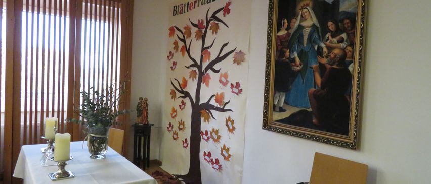 A room with a table, a candle, a plant, and a painting. A white cloth covers the table. A wooden chair with books is on the right. A large poster on the wall features a tree with autumn leaves.