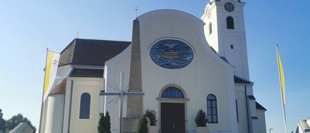 A white church with a steeple and clock stands under a clear blue sky. A cross and a flag are in front.
