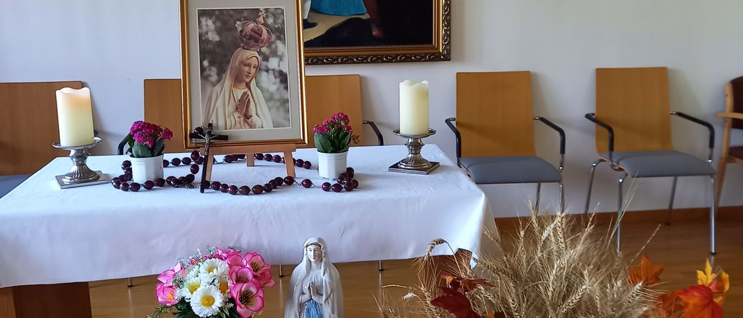 Ein kleiner Altar in einer Kirche mit einem gerahmten Bild der Jungfrau Maria, Blumen, Kerzen und einem Kreuz an der Wand.