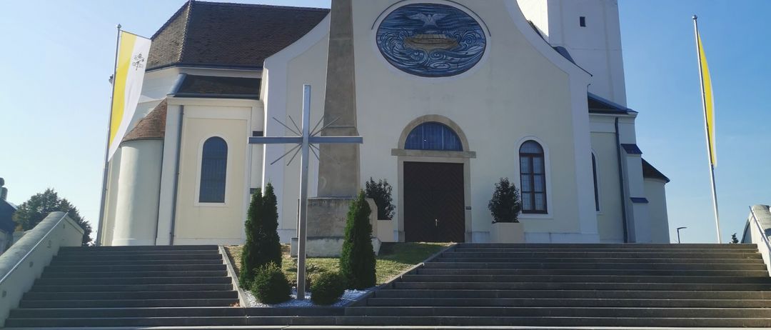 Eine weisse Kirche mit einem Turm und einem Kreuz davor. Ein Kreuz auf einem Steinsockel steht vor dem Eingang. Die Kirche hat ein grosses Fenster über der Tür.