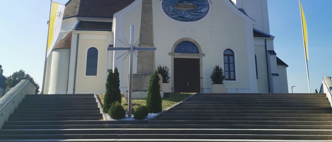 Eine weiße Kirche mit einem Turm, einer Uhr und einem Kreuz davor. Zwei Fahnen an Stangen stehen an den Seiten. Eine Statue steht vor dem Eingang.